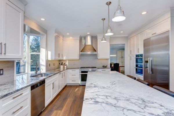 Luxury home interior boasts amazing white kitchen with custom white shaker cabinets, endless marble topped kitchen island and stainless steel appliances over wide planked hardwood floor.